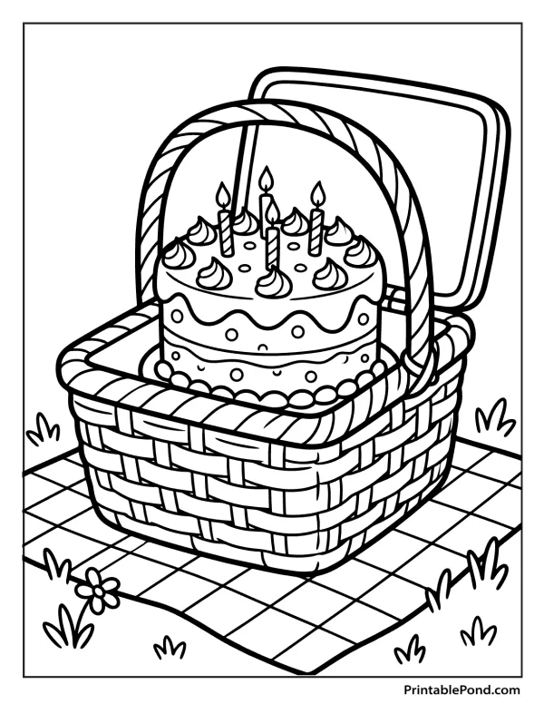 A two-tiered birthday cake with lit candles inside a woven picnic basket on a checkered blanket.