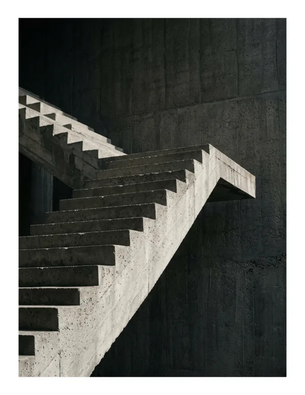 A brutalist concrete staircase with harsh lines ascends into darkness against a textured concrete wall.