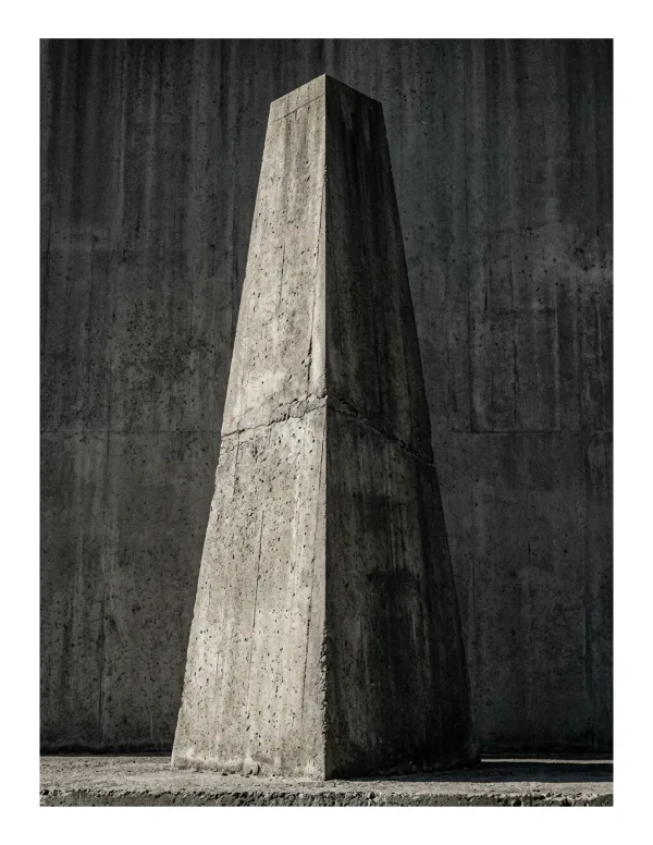 A tall, tapering concrete monolith stands in front of a rough concrete wall.
